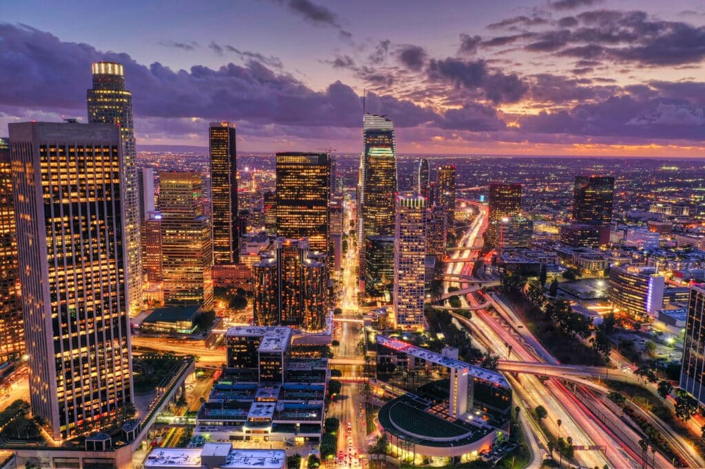 Aerial view of a vibrant city at dusk, showcasing illuminated skyscrapers, busy highways, and a colorful sunset sky.