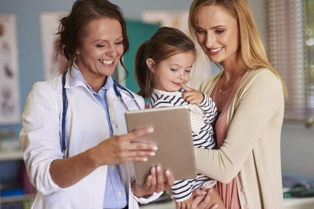A pediatrician shows a tablet to a young girl who is held by her mother in a medical office, all smiling.