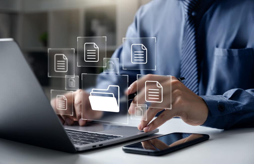 A person in a blue shirt working on a laptop with digital file icons floating above the screen, suggesting file management or cloud computing, with a smartphone beside the laptop.