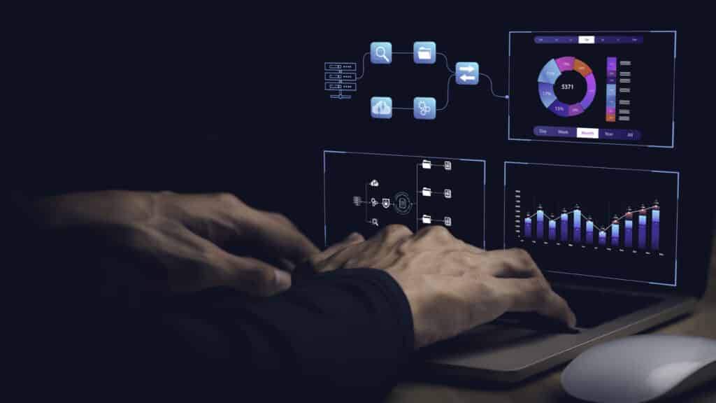 Hands typing on a laptop with digital analytics and data visualization graphics displayed on the screen, in a dimly lit room.