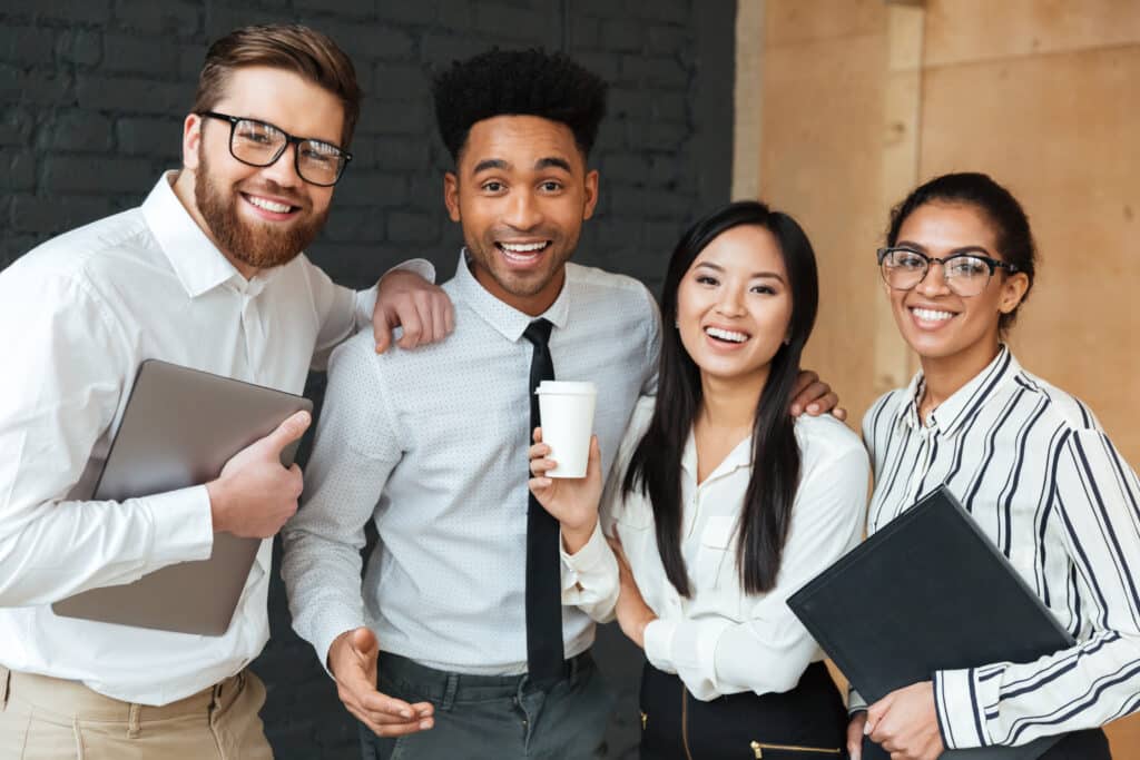 Four diverse professionals smiling and posing together in a modern office, holding documents and a coffee cup.