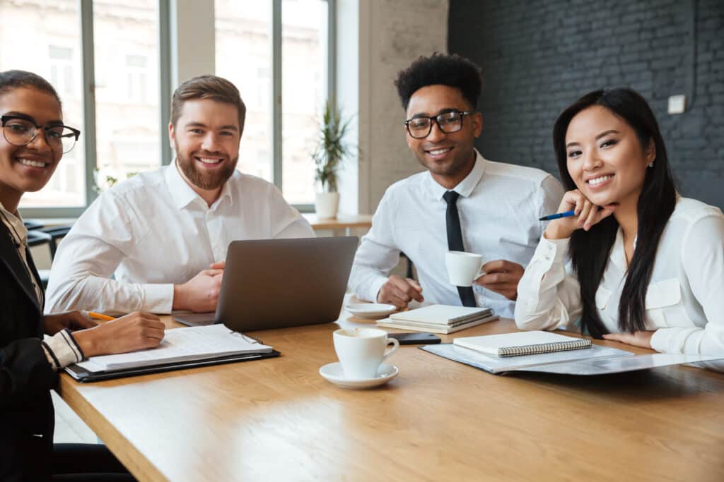 Four professionals smiling at the camera, seated around a table with a laptop, notebooks, and coffee mugs in an office setting.