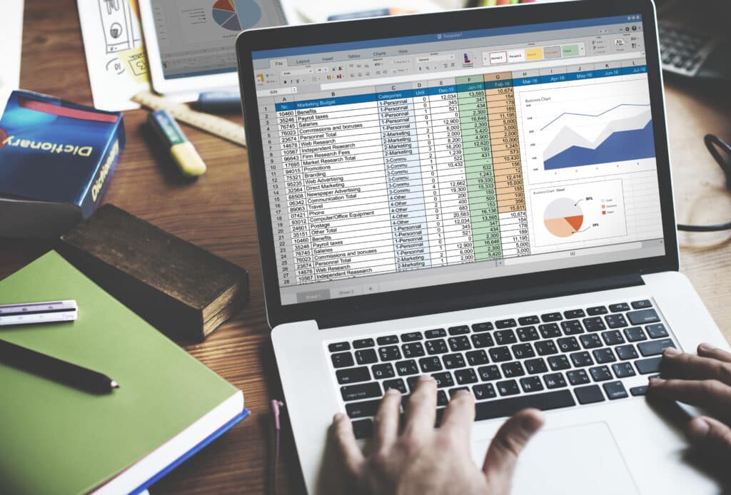 Hands typing on a laptop displaying detailed financial spreadsheets and graphs, surrounded by books and notes on a wooden desk.