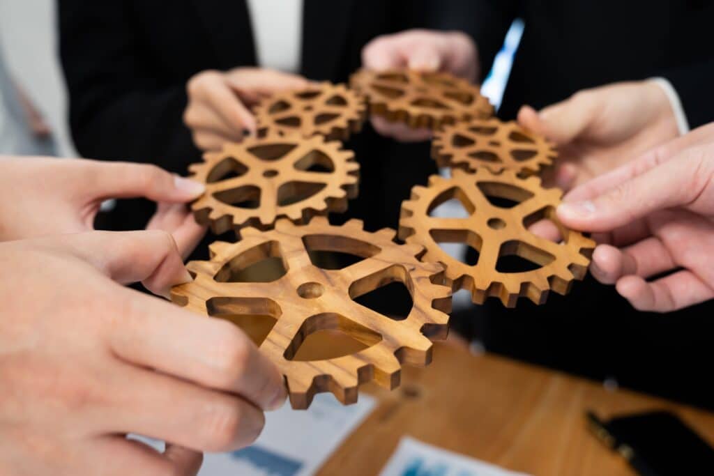 Four hands holding and connecting wooden gears over a table, symbolizing teamwork and collaboration.