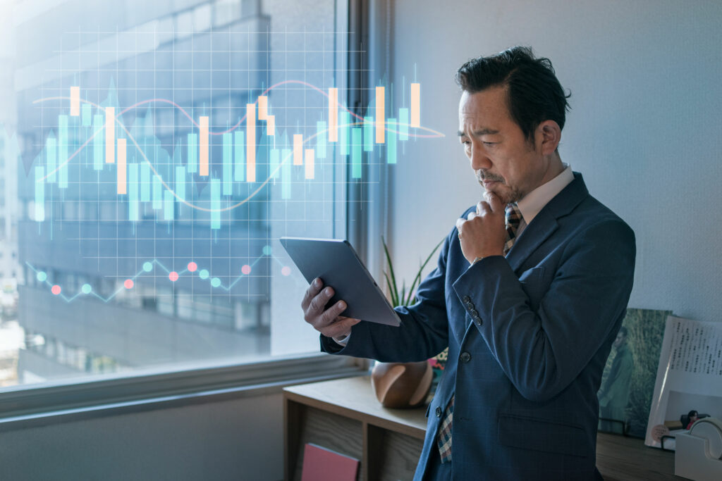 A businessman analyzes financial graphs on a tablet, standing by a window in an office.