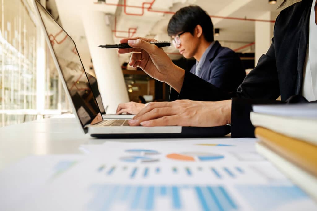 Two professionals analyzing data on a laptop with documents and charts on the table in a modern office setting.