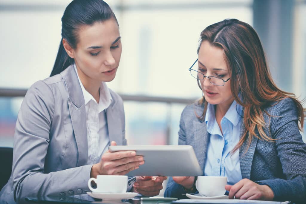 Two businesswomen reviewing information on a tablet while sitting at a cafe table with coffee cups.