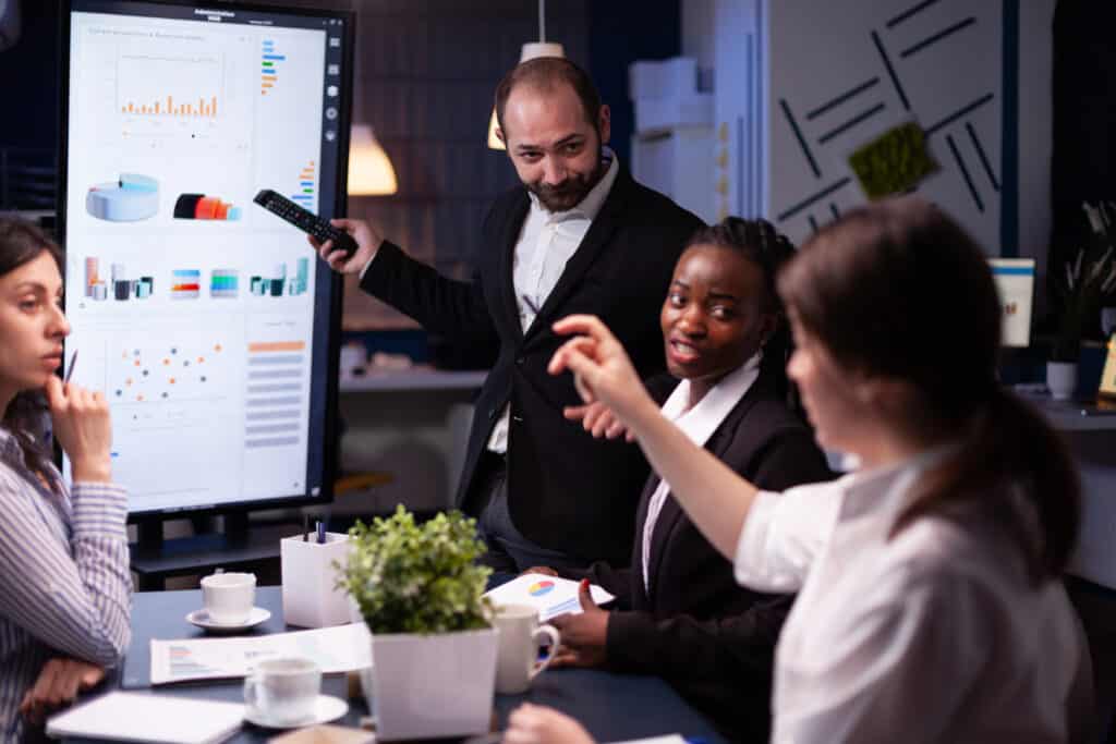 A diverse group of business professionals engaged in a discussion, with one man pointing at a digital screen displaying graphs during a night meeting in an office.