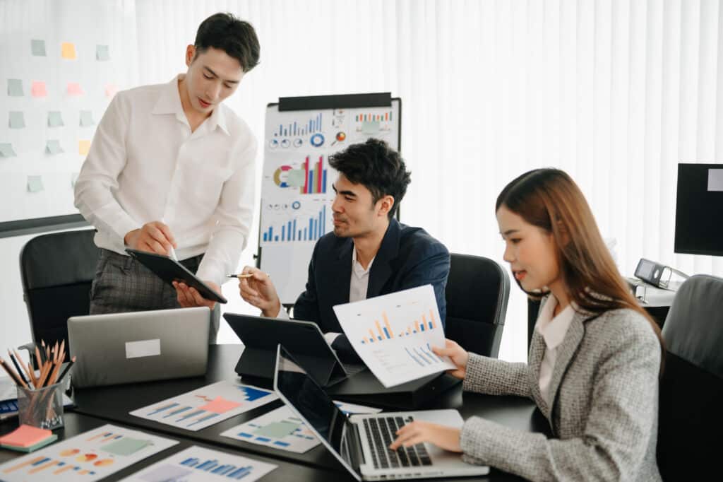 Three office colleagues discussing data charts on laptops and printed documents at a desk in a modern office setting.