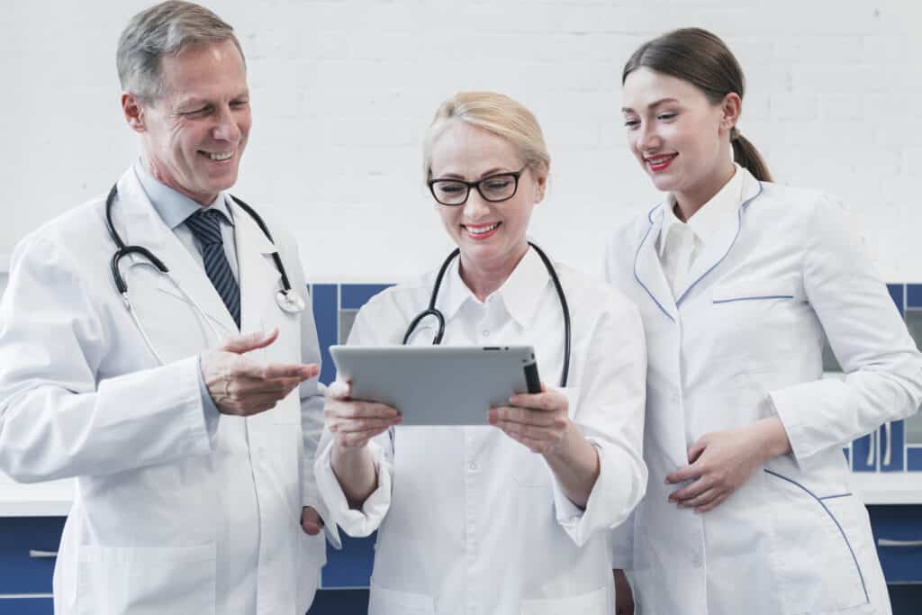 Three doctors, two women and one man, wearing white coats and stethoscopes, looking at a digital tablet and discussing in a hospital setting.
