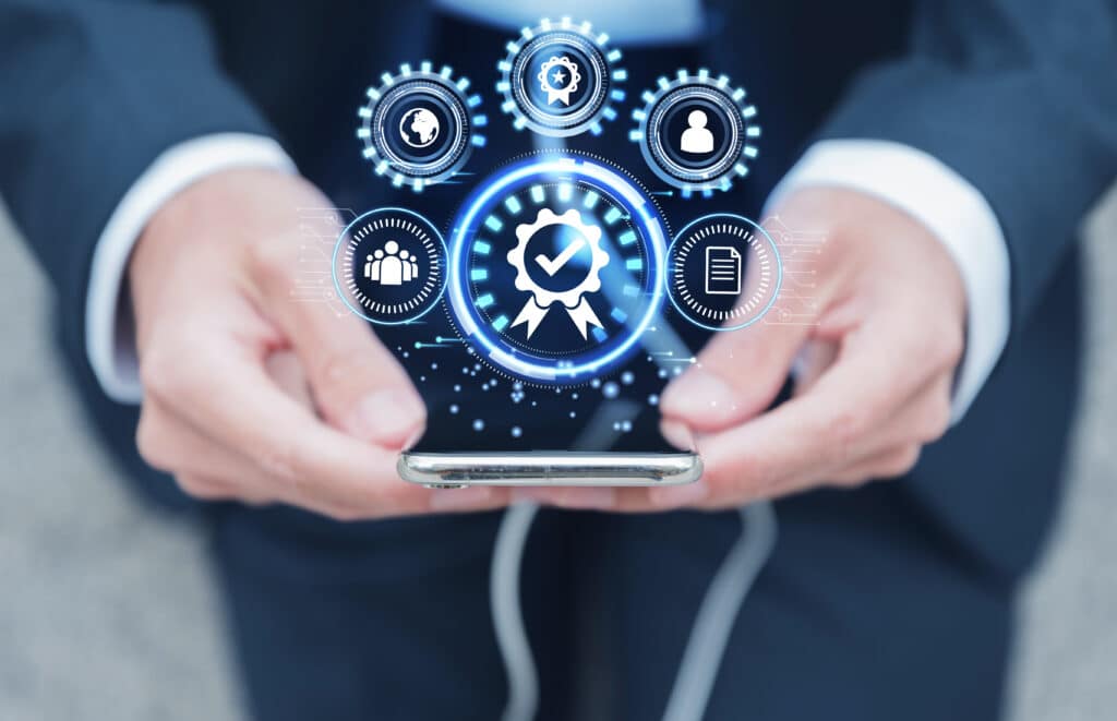 A businessperson holds a smartphone displaying holographic icons of gears and a central quality assurance symbol, symbolizing advanced technological management.