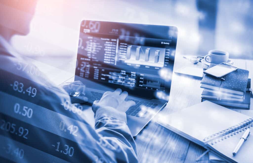 A person analyzing financial data on a laptop with stock market numbers overlay, sitting at a desk with notes and a coffee cup.
