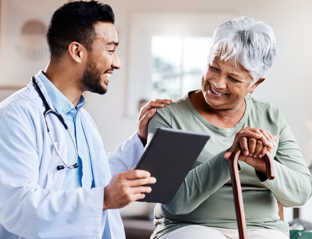Doctor showing a tablet to an elderly woman with a cane, both smiling in a cheerful medical consultation room.
