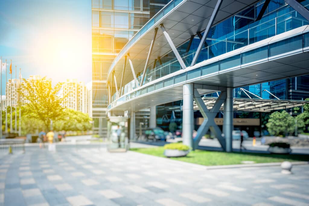 Modern glass office building with a steel structure entrance, bathed in sunlight, and adjacent to a plaza with trees, in an urban setting.
